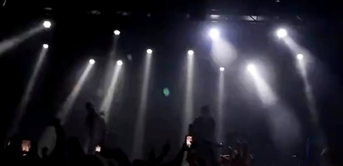 Concert stage lighting with multiple white beams over a dark crowd.